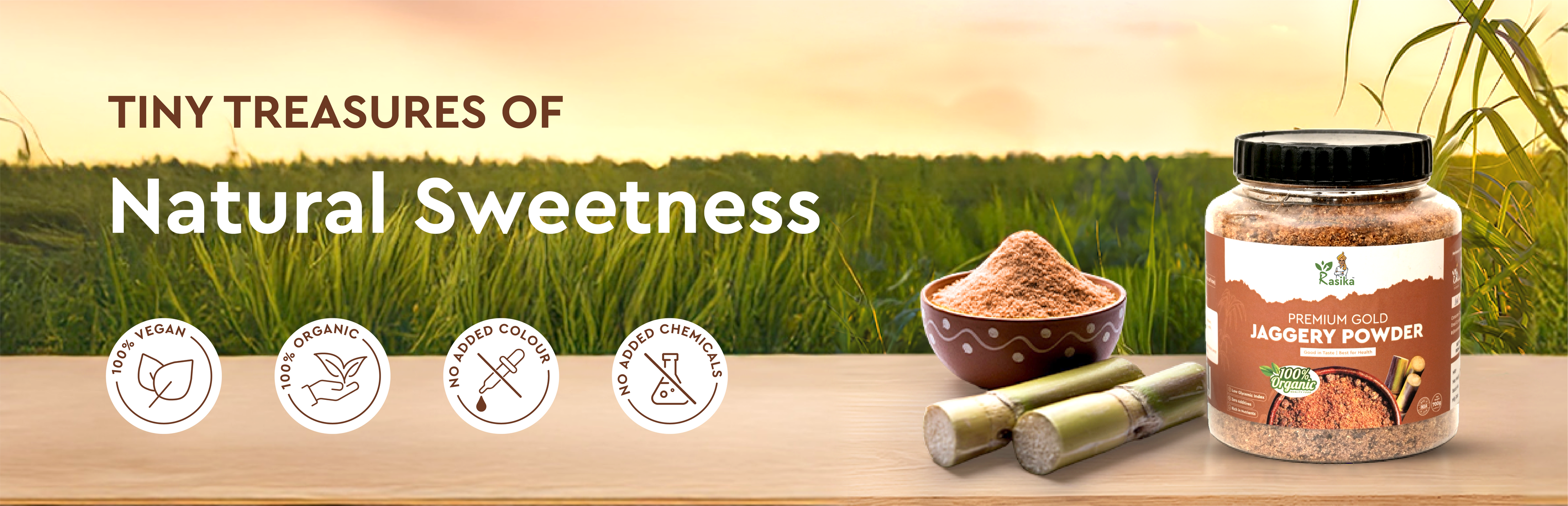 Jar of Rasika jaggery powder with a bowl of jaggery and sugar cane on a wooden surface with a grassy field background.